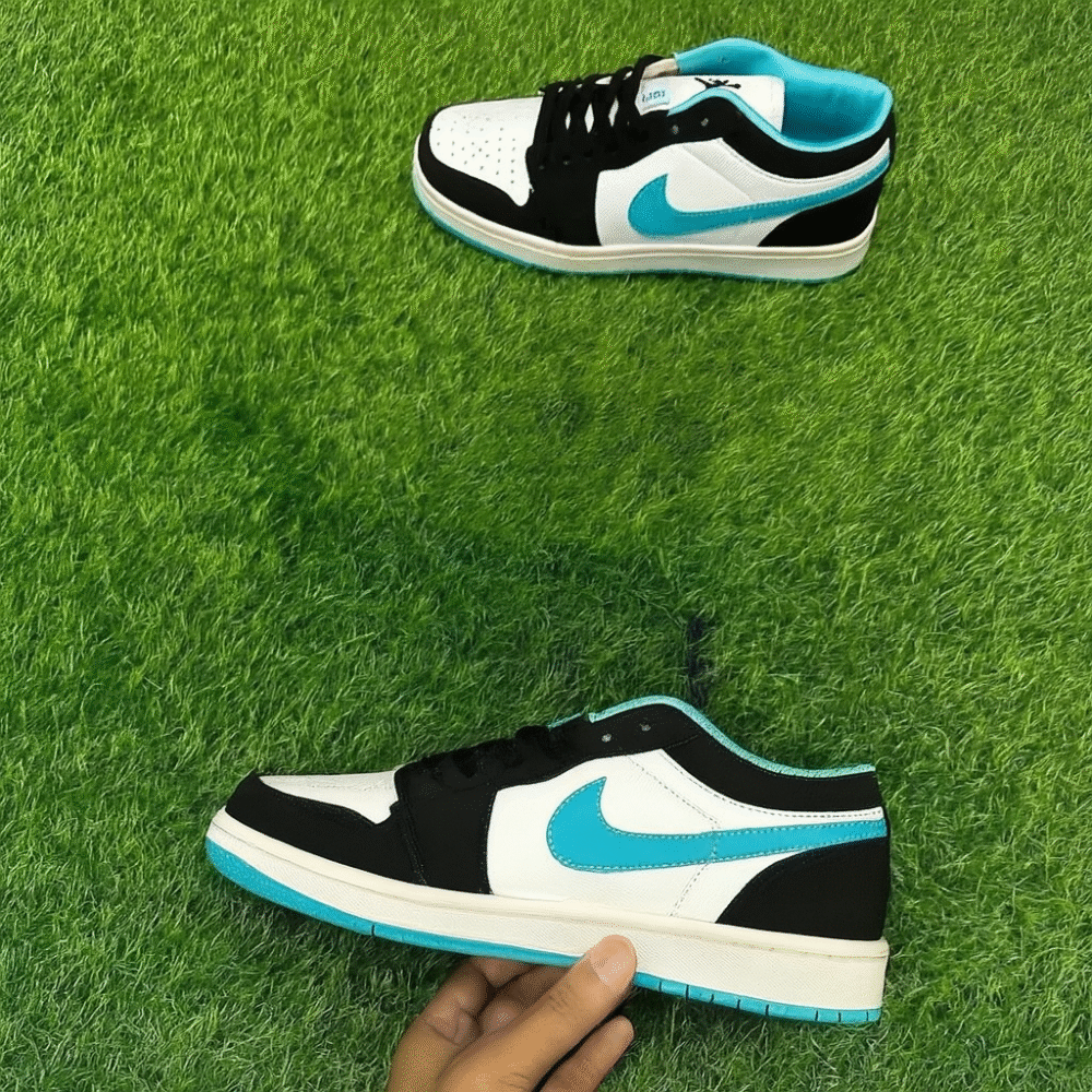 A stylish pair of black, white, and cyan low-top sneakers displayed on bright green artificial grass, with one shoe held in hand and the other laid flat.