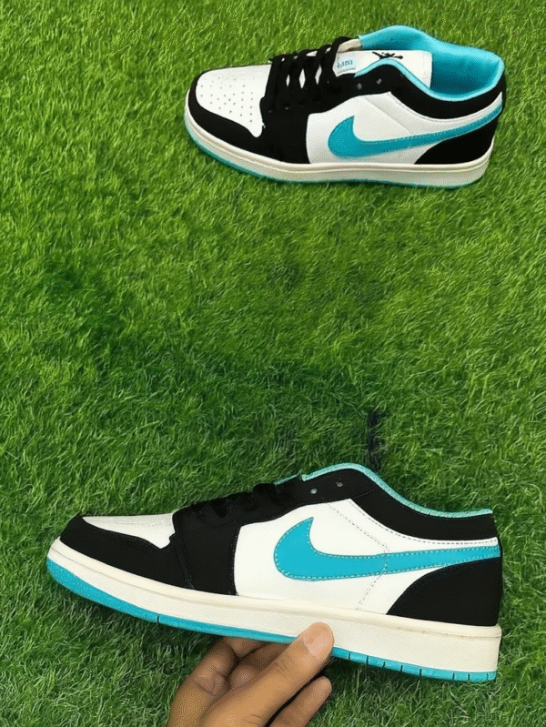A stylish pair of black, white, and cyan low-top sneakers displayed on bright green artificial grass, with one shoe held in hand and the other laid flat.