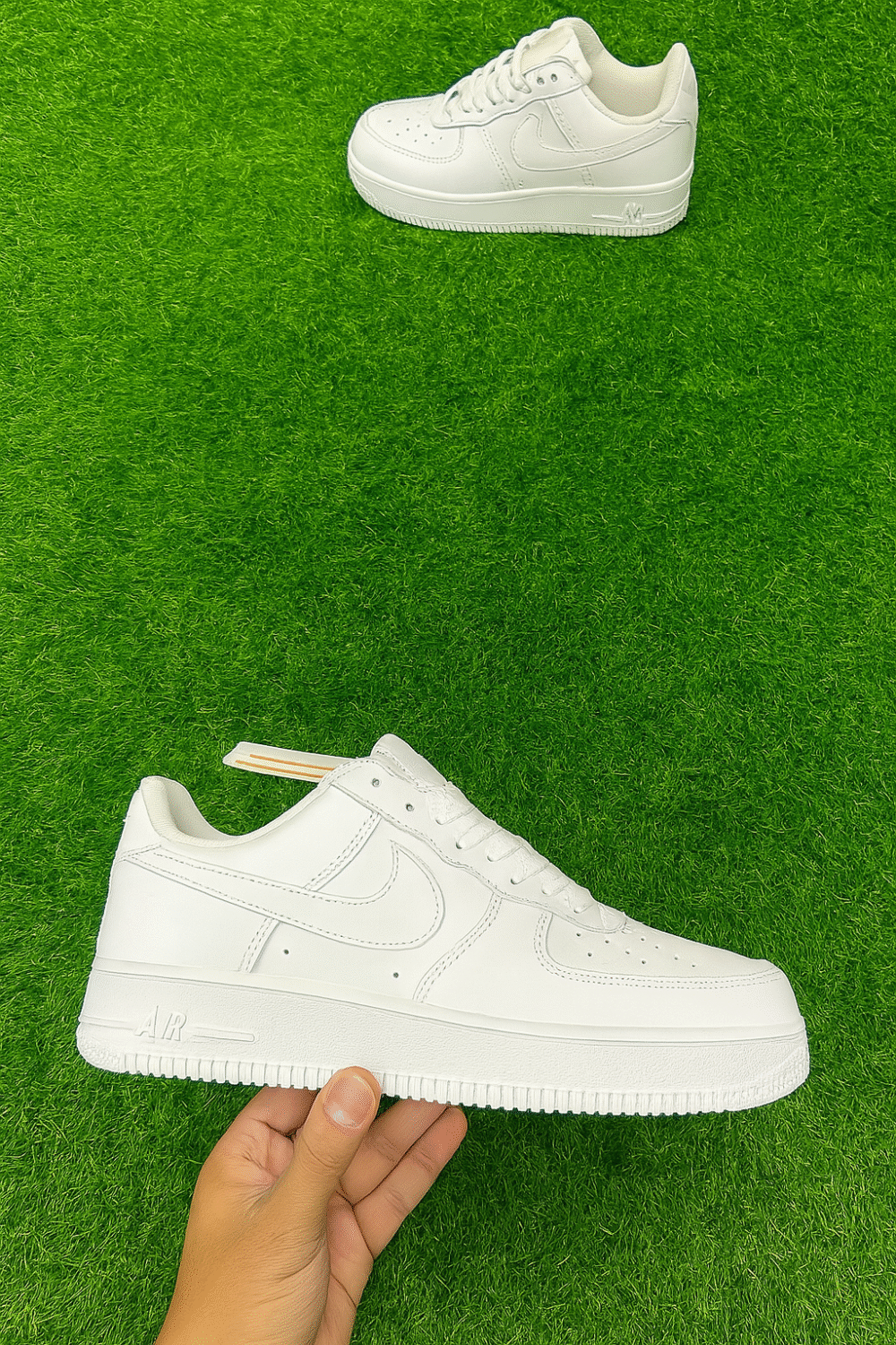 A pair of classic all-white Nike-style sneakers displayed on a vibrant green artificial grass surface, with one shoe held in hand and the other positioned above.