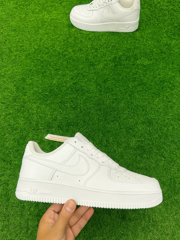A pair of classic all-white Nike-style sneakers displayed on a vibrant green artificial grass surface, with one shoe held in hand and the other positioned above.