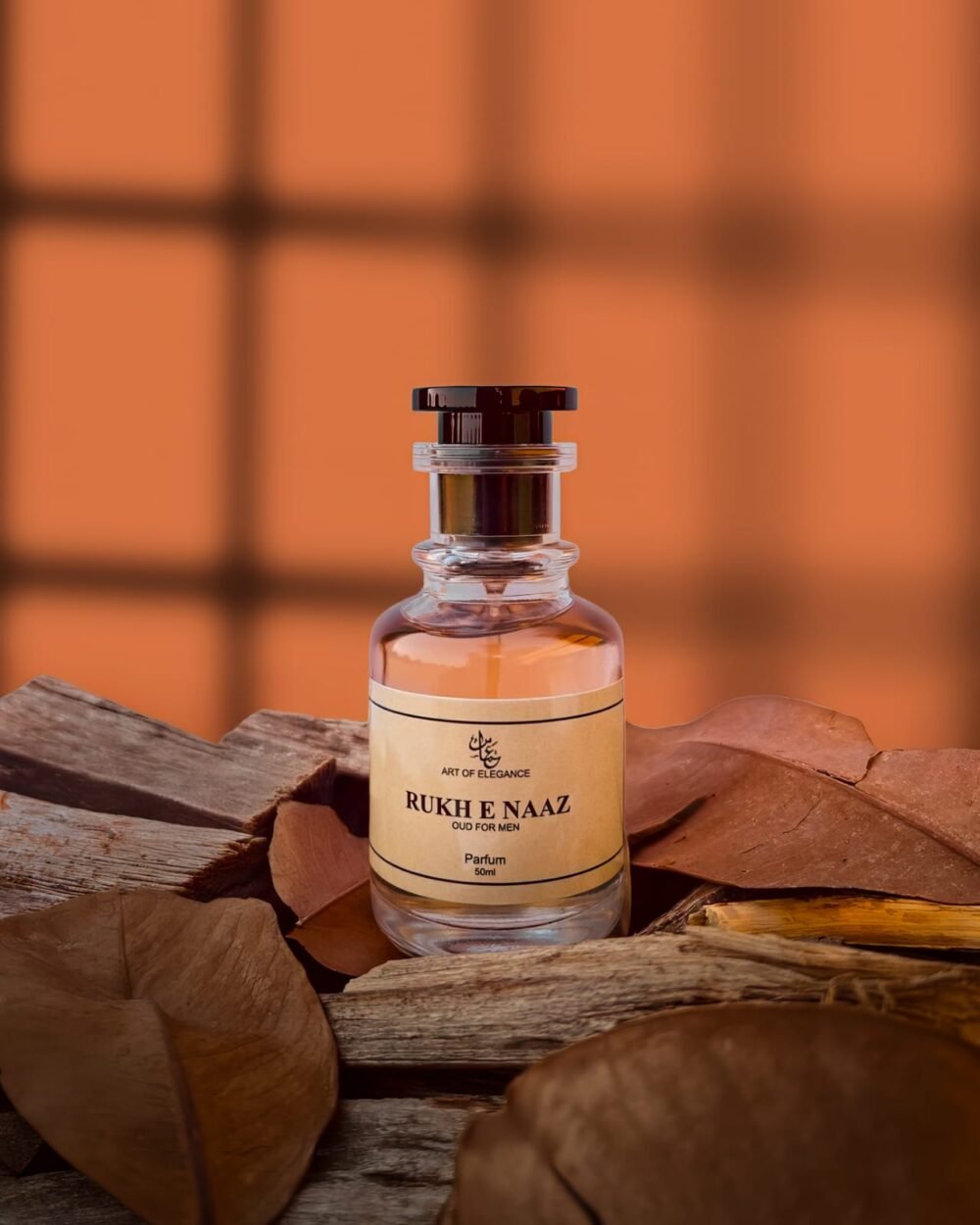 A premium glass bottle of “Rukh e Naaz – Oud for Men” perfume placed on rustic wooden planks with dried brown leaves, against a warm orange window-lit background.