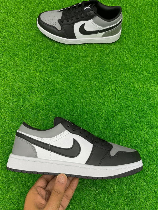 Black, white, and grey low-top sneakers displayed on vibrant artificial green grass, with one shoe lying flat and the other held in hand to showcase design details.
