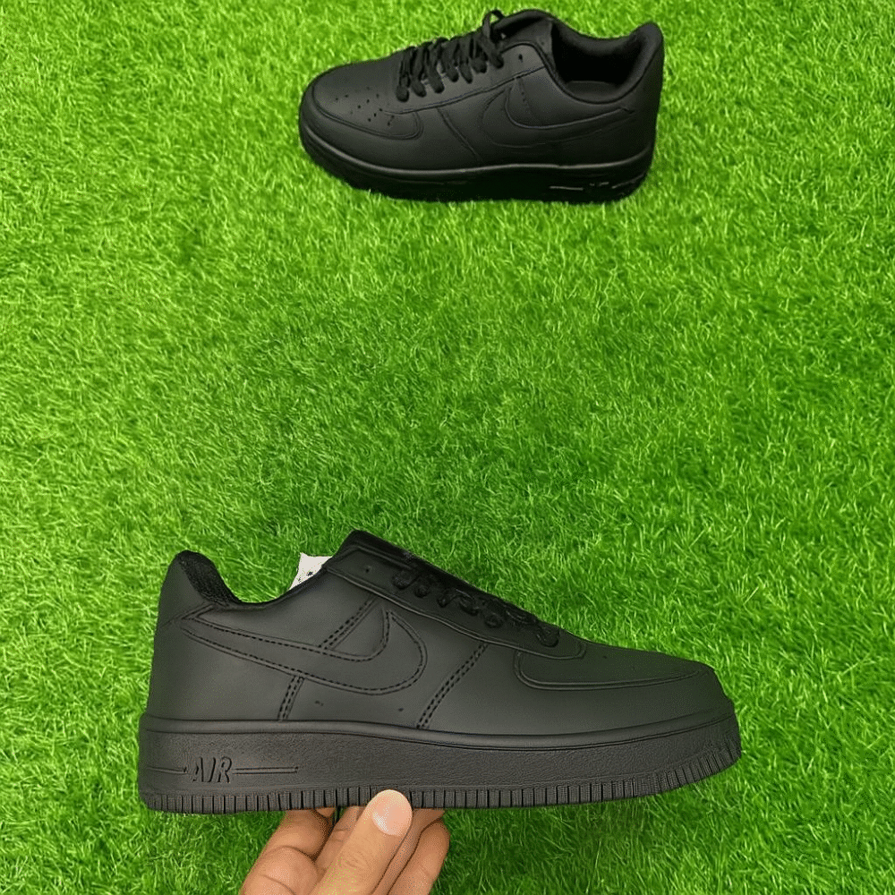A pair of all-black low-top sneakers placed on artificial green grass, with one shoe held in hand and the other positioned above, showing clean, minimal detailing.