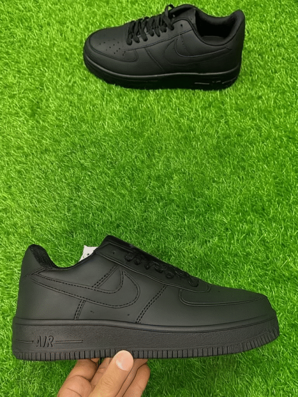 A pair of all-black low-top sneakers placed on artificial green grass, with one shoe held in hand and the other positioned above, showing clean, minimal detailing.