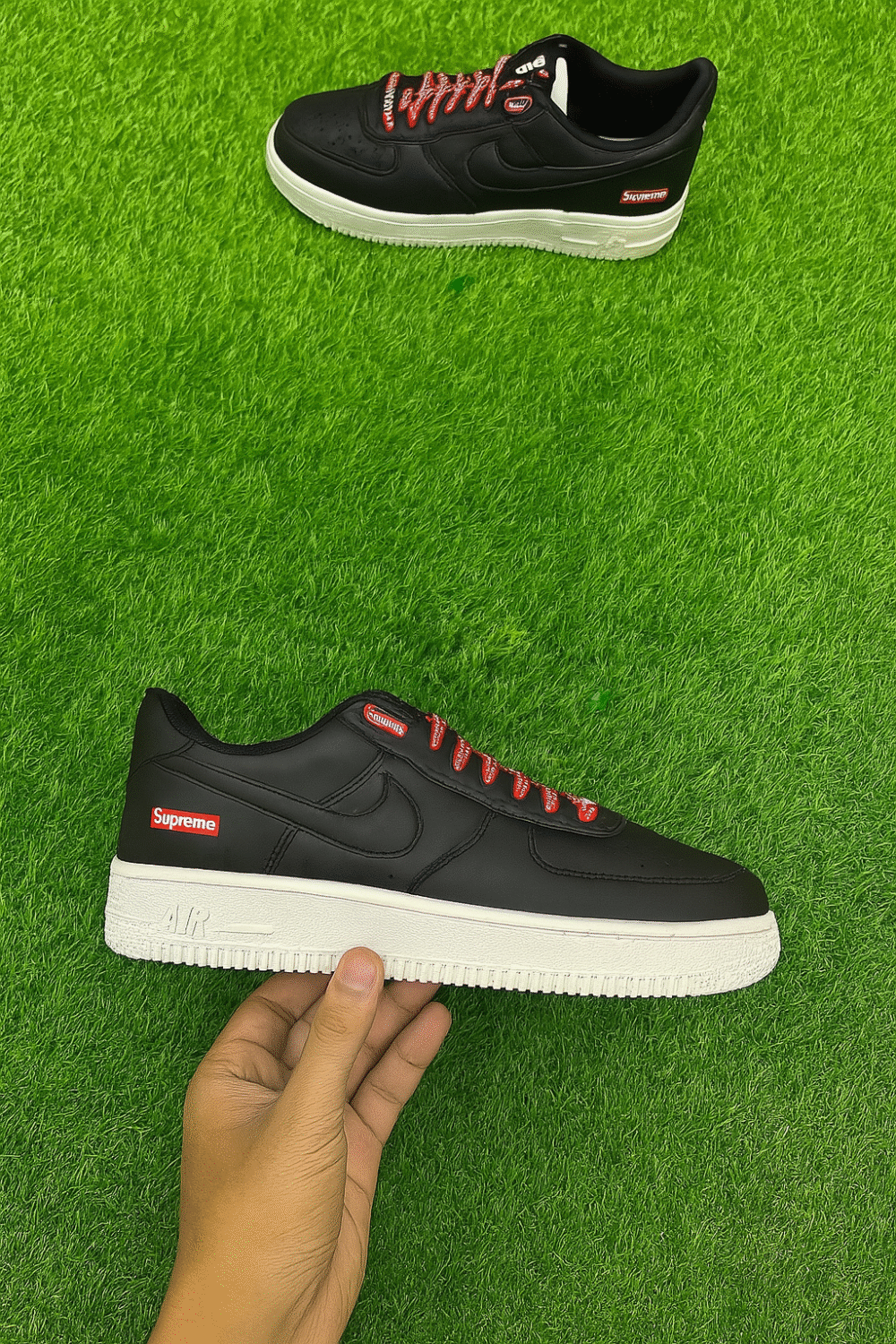 A pair of black Supreme-branded low-top sneakers with red laces and a white sole, placed on vibrant artificial green grass, with one shoe held in hand.