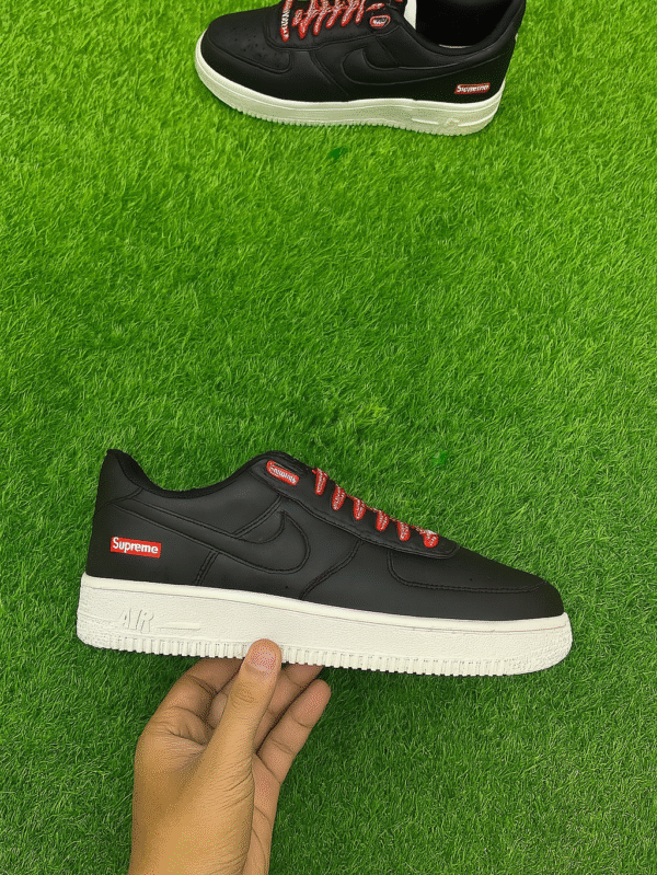 A pair of black Supreme-branded low-top sneakers with red laces and a white sole, placed on vibrant artificial green grass, with one shoe held in hand.